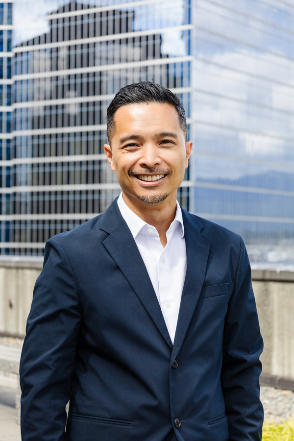 Team Portrait of Marc Bitanga, standing in a dark blue suit in front of a glass office building.